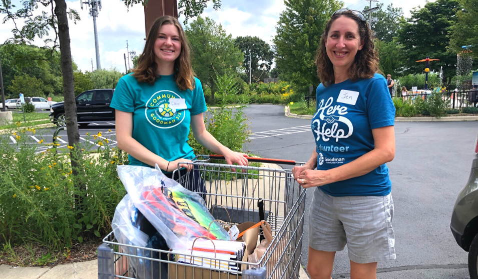 Volunteers wearing Goodman Center t-shirts stand with a shopping cart filled with school supplies.