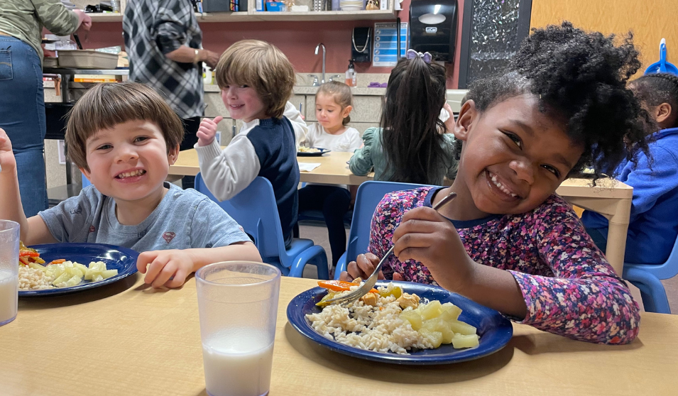 Two preschool students smile at the camera. In front of them is a plate of