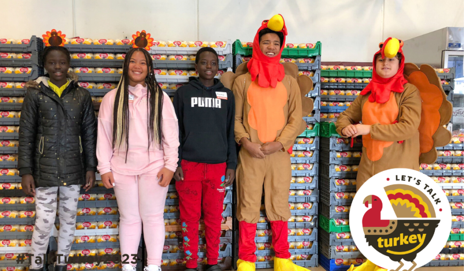 A group of teens smile in front of stacks of dinner rolls at the Goodman Center's annual Thanksgiving Basket Drive. Two of the teens are dressed in turkey costumes.