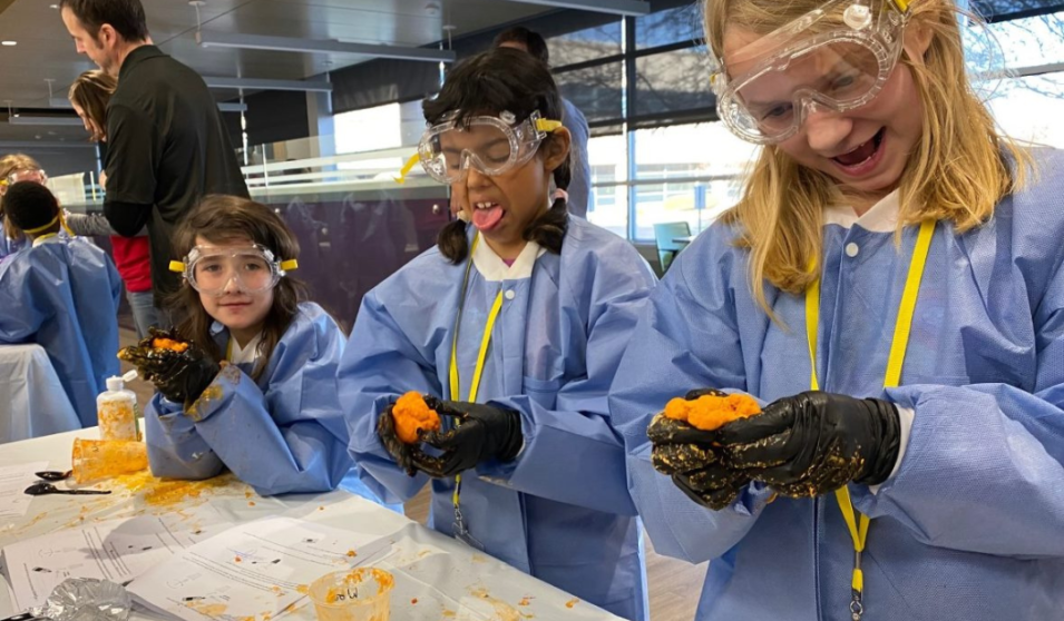 Three young girls wear lab goggles and gloves, holding homemade slime. One girl sticks out her tongue.