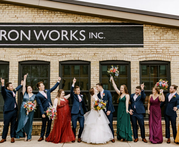 A wedding party in colorful dresses holding colorful flowers, posing and celebrating in front of Goodman's Ironworks Building
