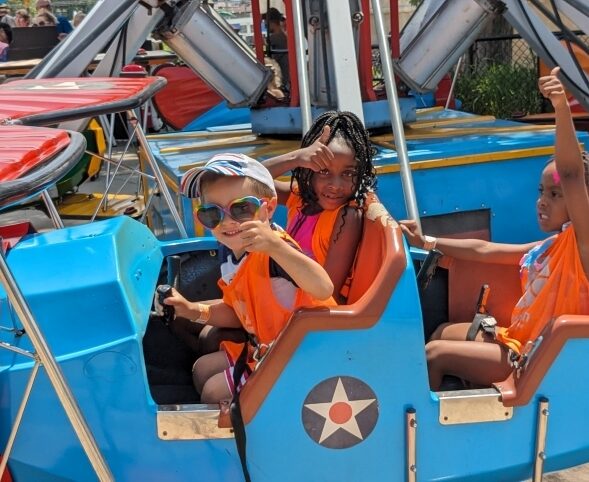 Three elementary schoolers giving thumbs up as they wait for the start of a Mt. Olympus park ride. Two are sitting next to each other in the front seat of the blue ride car, while one sits by herself in the second row.