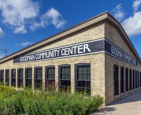 A photo of the Goodman Center's Ironworks building, taken from Waubesa Street. The center's name is painted on the side of the building in block letters, and there are wildflowers growing in the foreground.