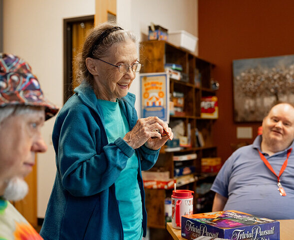 Three older adults playing Trivial Pursuit; laughing woman reads from card while two men contemplate answer
