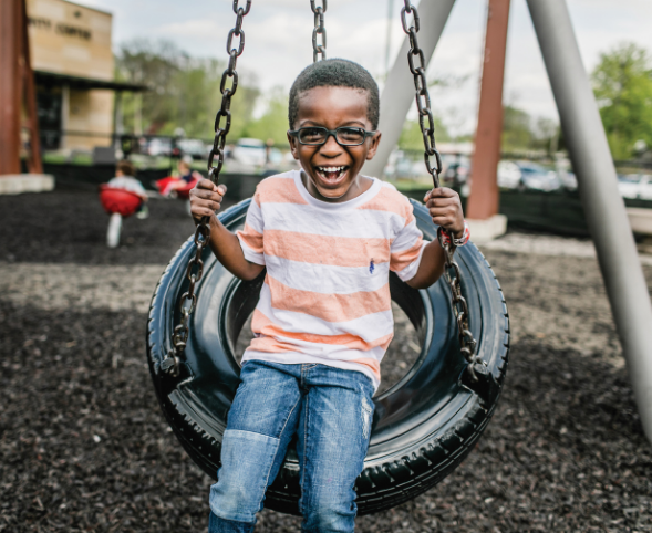 An African American child wears a striped shirt and glasses and smiles while swinging on a tire swing.