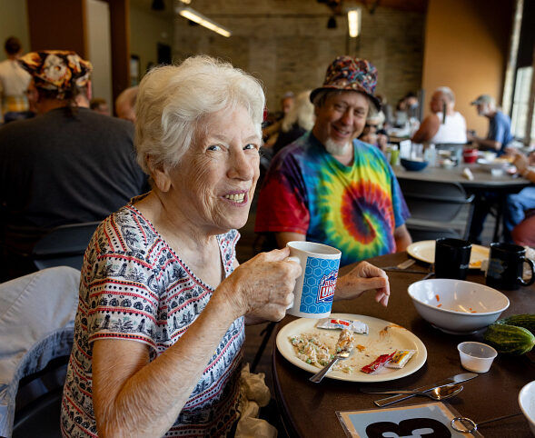 An older woman smiles at the camera holding a coffee mug. A man in a rainbow shirt and colorful bucket hat laughs happily behind her.