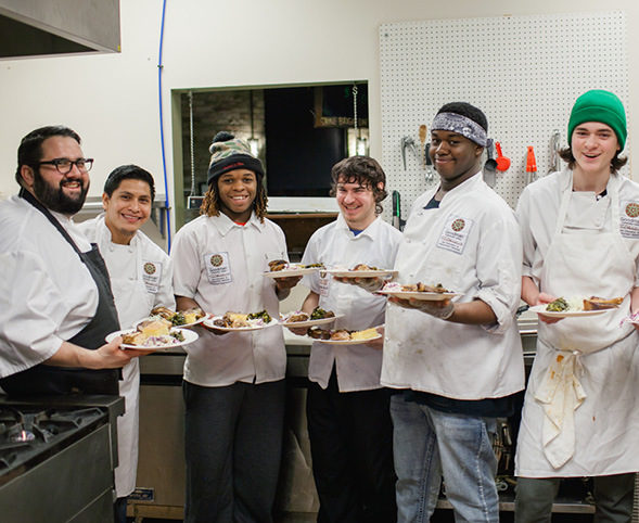 A group of teens and a chef in a professional kitchen holding plates