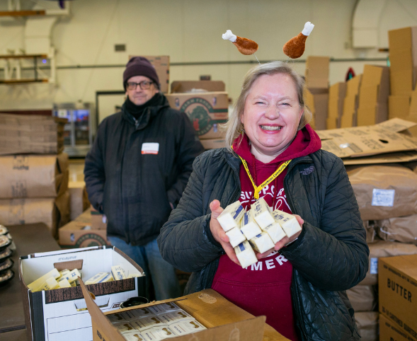 A woman smiles at the camera as she holds up sticks of butter and wears a turkey leg headband at the Goodman Center Thanksgiving Basket Drive.