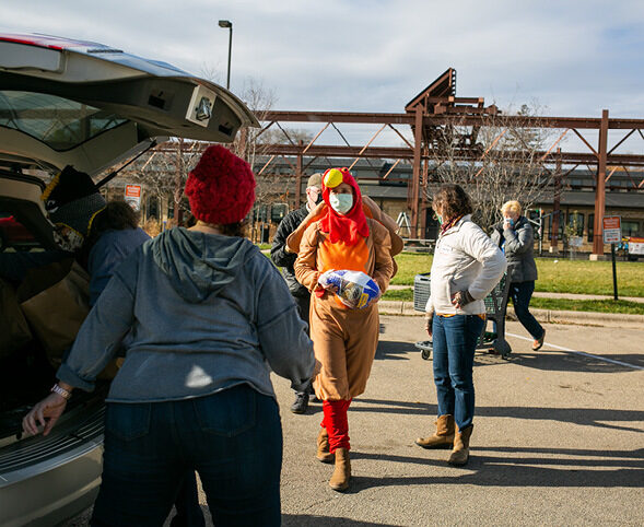 A person in a turkey costume delivering a frozen turkey to a parked car, with volunteers helping