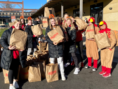 A group of UW Madison athletes hold grocery bags and smile at the Goodman Center's Thanksgiving Basket Drive. Two athletes are in turkey costumes.