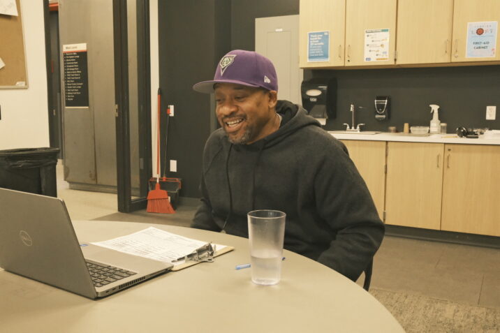Arthur Morgan is sitting in front of a round table in an unseen chair. He has his laptop open and is answering an interview question while doing some paperwork. There's a glass of water to the right of the laptop. Arthur is leaning towards the table and is wearing a black hoodie and purple baseball cap with the lid facing forward. A clipboard is placed horizontally on the table between Arthur and the laptop. Arthur is mid-sentence and looks happy as he continues answering a question.