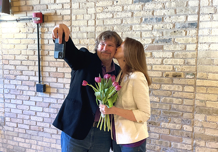 A photo of a couple taking a selfie. She is kissing his cheek while holding a bouquet of tulips.