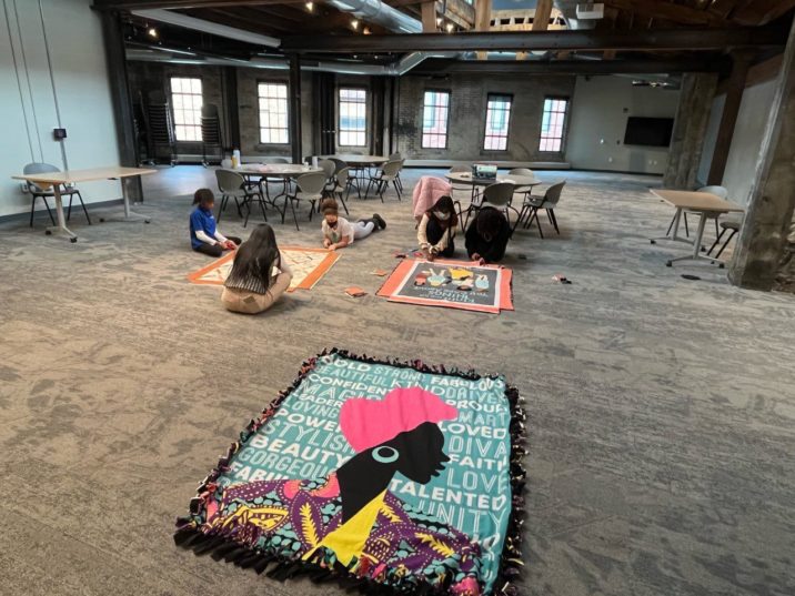 In the foreground, a beautiful fleece blanket featuring the silhouette of an African woman. In the background, girls work on making blankets.