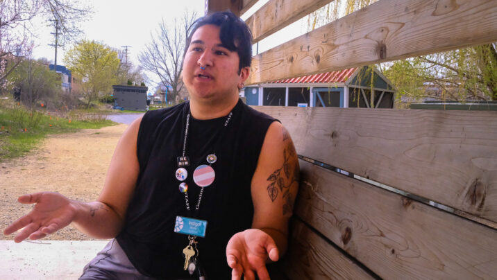 Indigo Alcorta is sitting on a brown wooden bench outside. His hands are casually placed in front of him palms up, and he is using them to emphasize what he is saying. Inigo is wearing a black tanktop and his teal Goodman name tag and key hang from a black lanyard. On the lanyard are many buttons, including a transgender pride flag button, a he/him pronoun button and a rainbow button. Indigo is looking off to the left as he gives an answer to a question, looking pensive while mid-sentence.