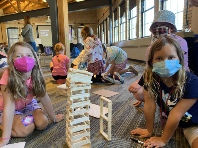 Three girls smile behind masks with a tower made out of balsam wood between them.