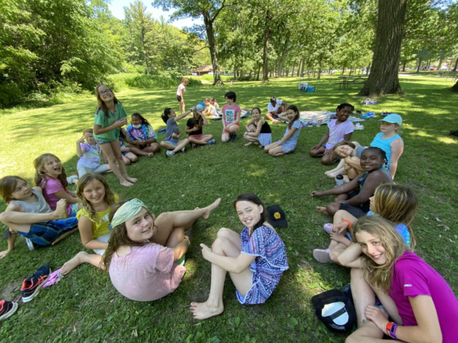 A large group of girls sit in a circle on the grass and smile for the camera.