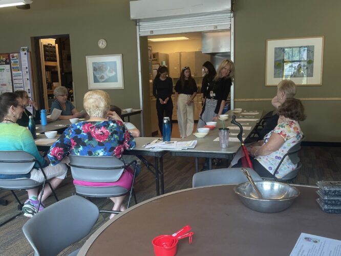 A group of people are sitting at a u-shaped table, listening to Older Adult Manager, Gayle, introduce the med students about to lead the class. Gayle and the med students are standing in the open kitchen entryway. The attendees have their backs turned to the camera and are waiting for instruction.