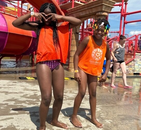 Two Black girls happily stand together at Mt. Olympus water park. Both are wet from a day of water park fun and are standing next to the park's pink and orange play structure in orange Goodman pinnies.