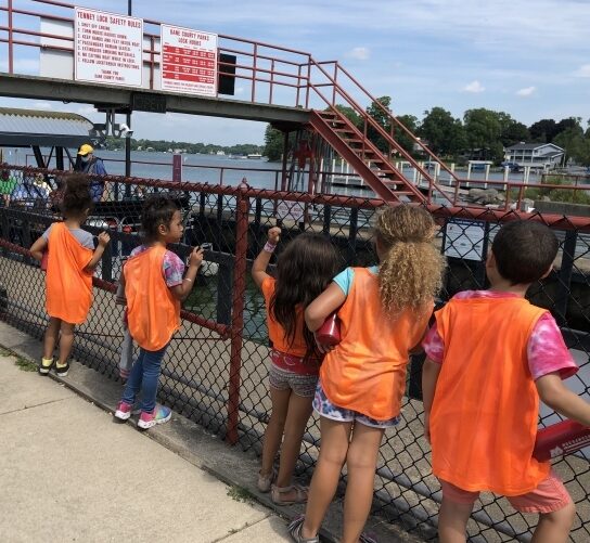 Five multiracial kids--four girls and a boy on the far right end--lean against a fence at Madison's Tenney park docs to look at the docking tools and watch a boat go out onto the lake. All of them are facing away from the camera with pure curiosity.