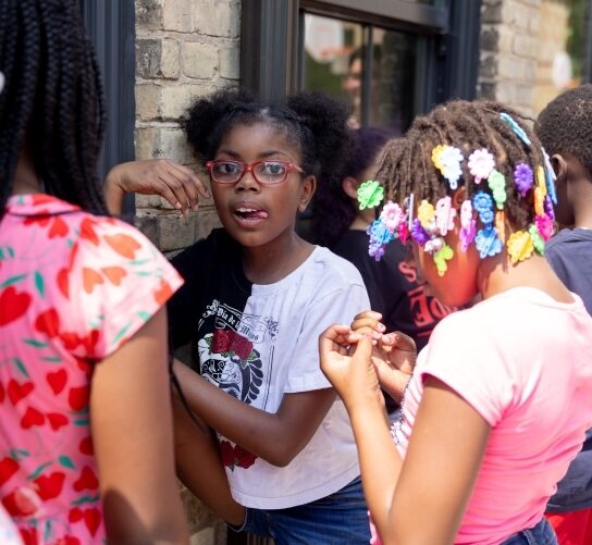 A late elementary age Black girl stands out amongst friends as they line up outside against the Ironworks building exterior. She's sticking her tongue out at the camera with one hand up, wrist bent at a 90% angle towards her face. Her hair is in two poms and she's wearing a white t-shirt with a red and black design. It's a sunny day and the rays shine on the girl to her right with colorful hair clips and a pink t-shirt shirt.