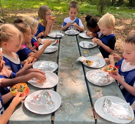 A group of multi-racial children wearing blue Goodman pinnies sit at a picnic table at Troy Gardens eating a snack off of white paper plates. The photo is taken from one end of the table, and a white boy is sitting on the opposite side. The left bench has 6 kids sitting together while the right bench has only four (one boy is hidden behind the head of another, but his plate is visible). The table is in the shade, but it is a sunny summer day, and the children are enjoying their food.