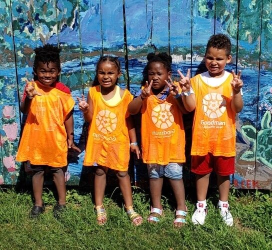 A group of four Black children stand posed together against a beautifully painted blue fence with a water scene. All four hold up piece signs, and are squinting as they face the sun to look at the camera. They're all wearing shorts and the orange Goodman pinnies.