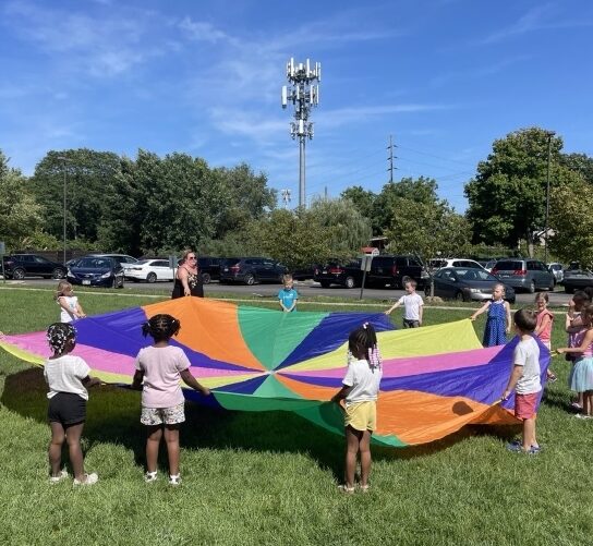 A group of multi-racial children stand outside together, each holding the edge of an outspread yellow, pink, blue, orange and green parachute. It's a sunny, clear blue day with just a few thin clouds scattered across the sky.