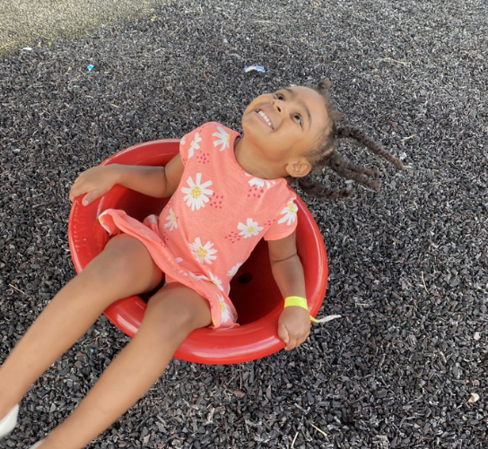 A smiling young girl lets her head lean back while she spins on a playground toy.