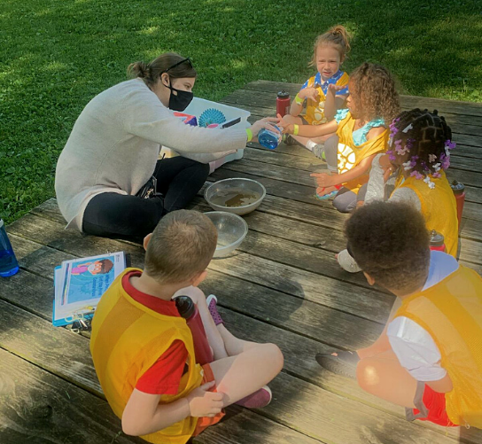 A preschool class sits in a circle, doing a science experiment with their teacher.
