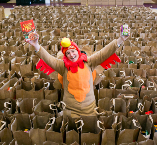 A person dressed in a turkey costume surrounded by bags packed for Goodman's Thanksgiving Basket Drive.