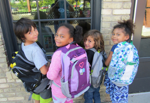 Four young kids stand with their backs to the camera while wearing backpacks. They look over their shoulders smiling.