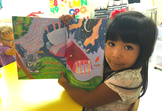 A preschool girl holds up a book she's reading to show the pictures inside.