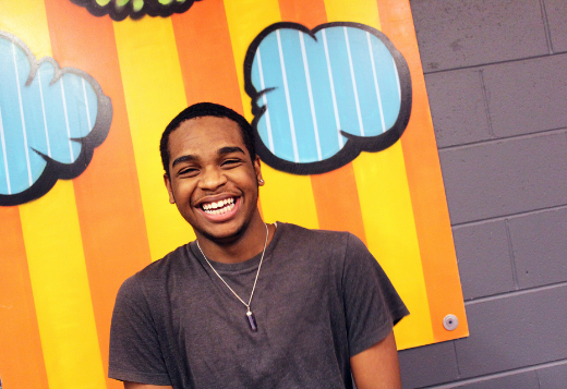 An African American teen smiles at the camera. Behind him is artwork in bright orange and yellow stripes.