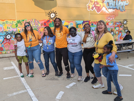 A group of staff, volunteers, parents and kids link arms and smile in front of a brightly colored abstract wall mural.