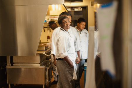 A teen looks over his shoulder and smiles from inside the Goodman Center's professional kitchen. He is dressed in his catering uniform.