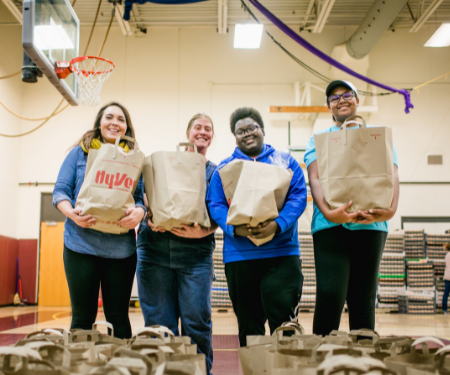 Four volunteers stand in the Goodman Center gym holding grocery bags and smiling at the camera.