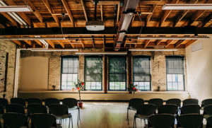 An industrial room with chairs set up to create an aisle, and two flower arrangements in front of sunny windows