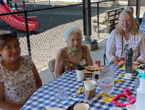3 women sit together at their picnic lunch table smiling at the camera.